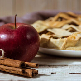 Im Hintergrund ein Apfelkuchen im Vordergrund ein Apfel und Zimtstangen